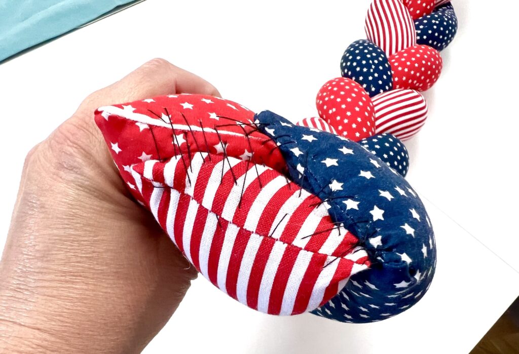 A hand holds a partially sewn, fabric necklace made of red, white, and blue patterned sections with visible black stitches.