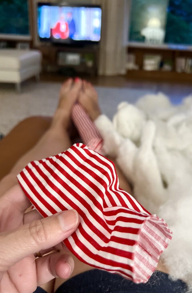 A person holds a red and white striped fabric piece while lying on a couch with stuffing on their legs; a TV is on in the background.