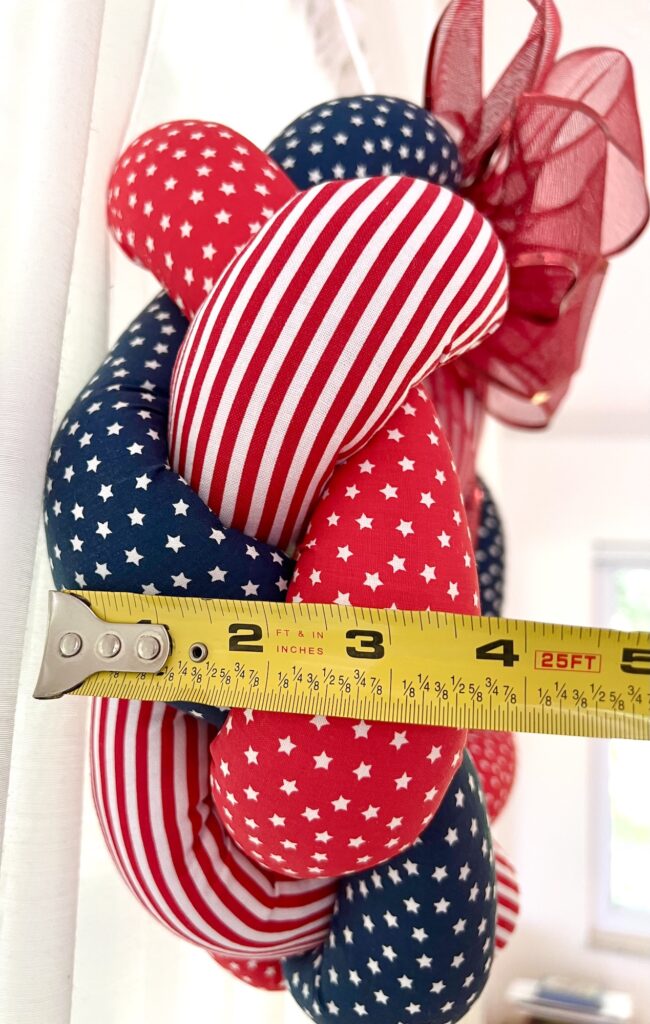 A tape measure shows the width of a red, white, and blue star-patterned wreath, with a window and red mesh bow visible in the background.