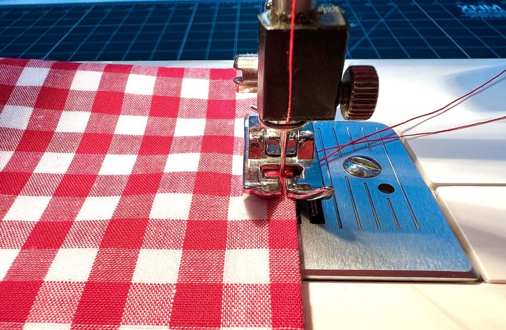Close-up of a sewing machine stitching red and white checkered fabric, with red thread running through the needle and presser foot.