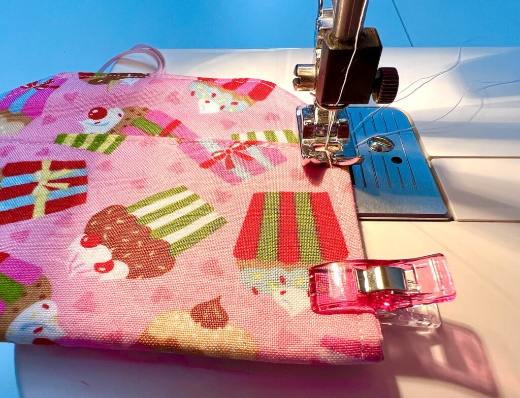 A sewing machine stitches a pink fabric with a cupcake pattern; a pink fabric clip holds the material in place near the presser foot.