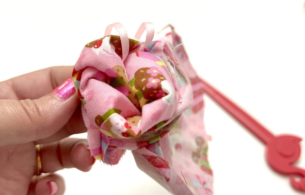 A hand holding a small pink fabric bundle with a mushroom pattern, partially gathered, against a white background.