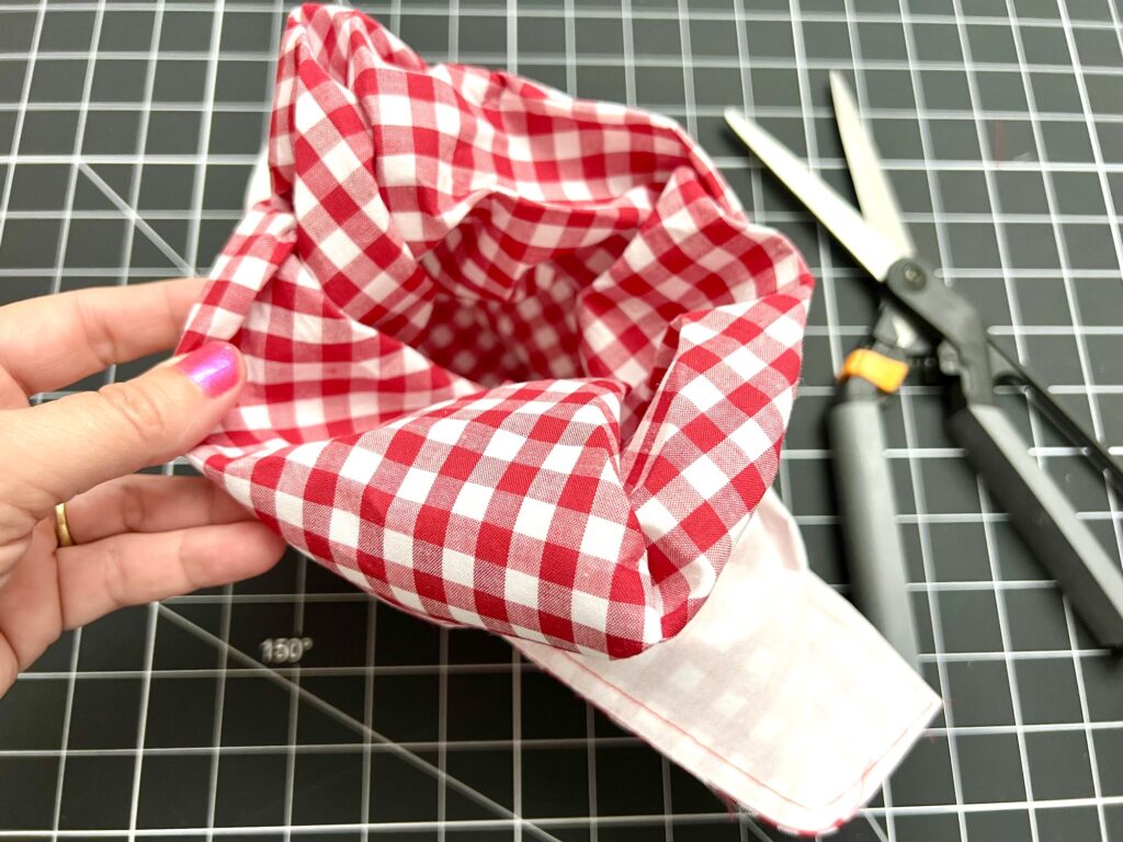 A hand holds an open red and white checkered fabric item on a cutting mat, with scissors and a rotary cutter nearby.
