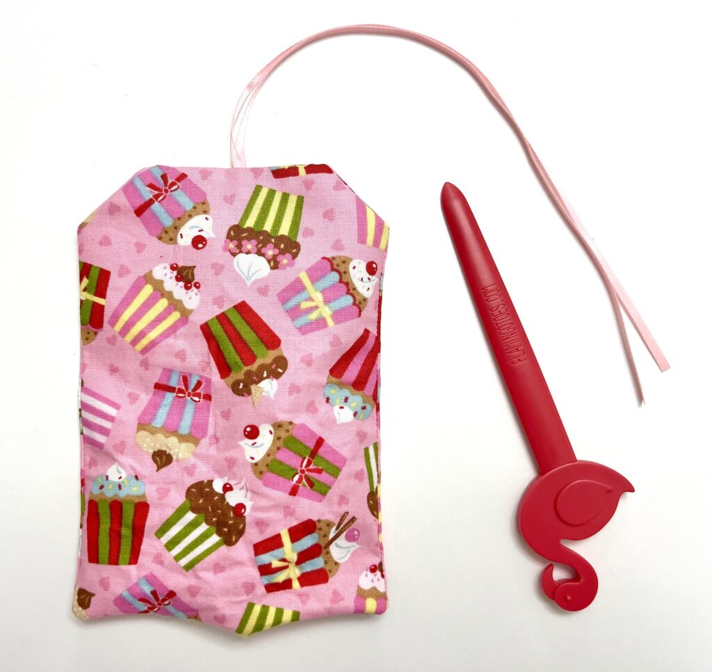 A pink fabric pouch with a cupcake pattern and a pink plastic tool with a hook shape, both placed on a white surface.