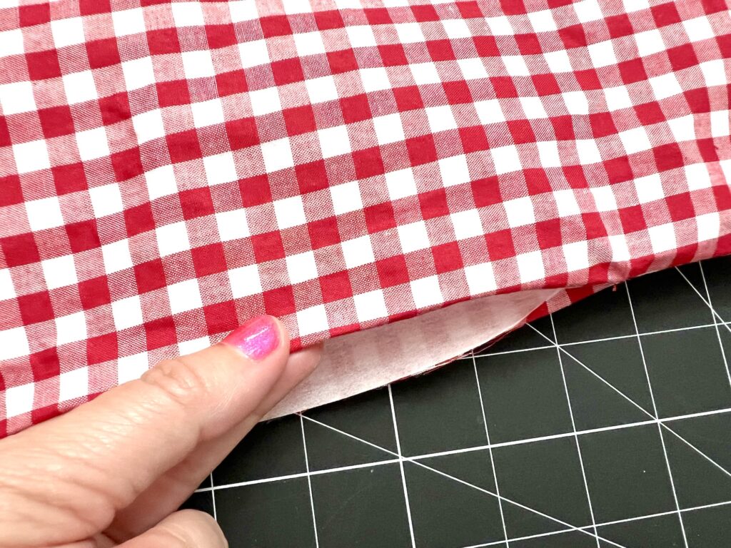 A hand with pink nail polish holds the edge of red and white checkered fabric, showing a partially finished hem on a black cutting mat.