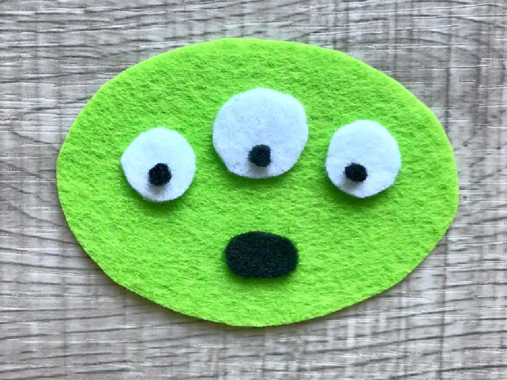 A green felt oval with three white eyes and a black mouth, arranged to resemble a surprised face, on a wood-textured background.