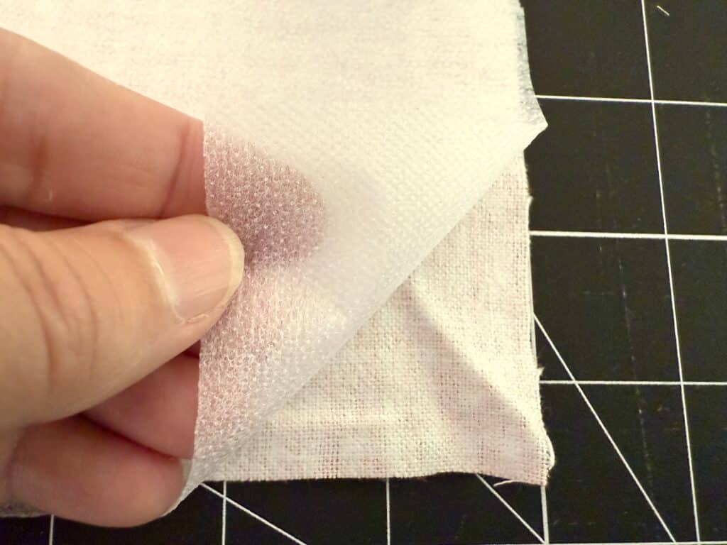 A hand lifts a sheet of white fusible interfacing from a square of fabric on a black cutting mat with white grid lines.