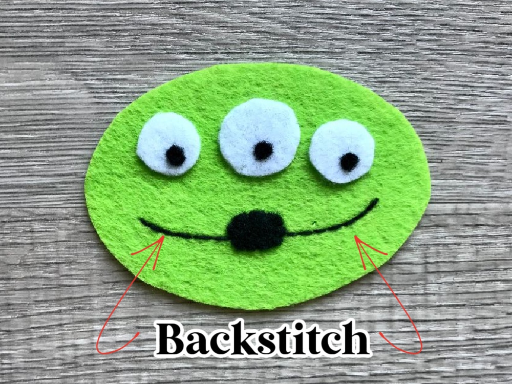 A green felt oval with three white and black eyes, a black nose, and a black stitched smile labeled "Backstitch" with red arrows pointing to the smile.