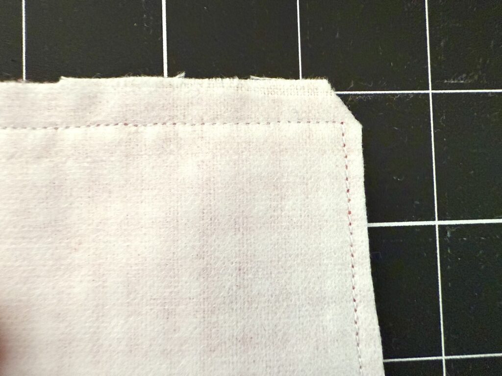 A crayon roll with notepad made of light-colored fabric and stitched edges is positioned on a black cutting mat marked with a white grid.