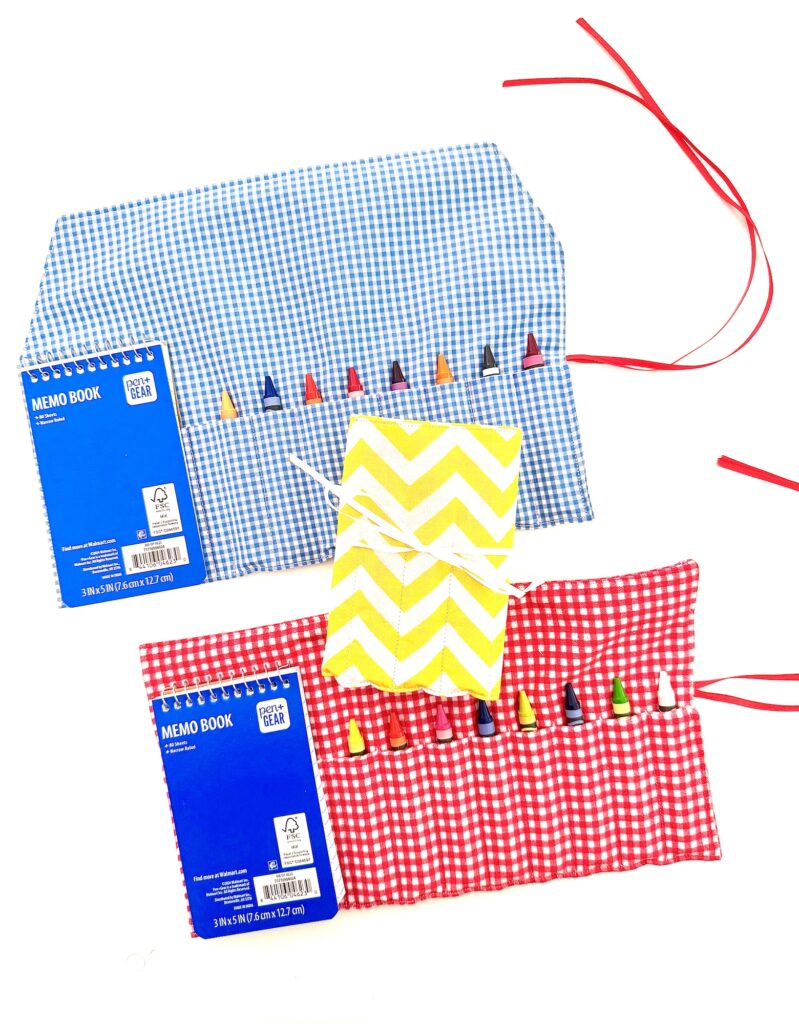 Two fabric pouches, one blue and one red, act as a crayon roll with notepad—each holds colored crayons and a memo book, while a yellow chevron-patterned notebook tied with string sits centered between them.