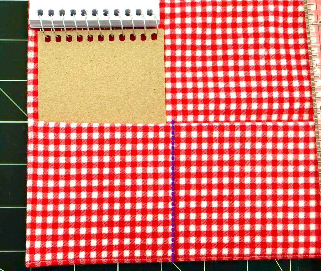 A red and white checkered crayon roll with notepad featuring four sections; one section holds a small spiral notebook with a brown cardboard cover.