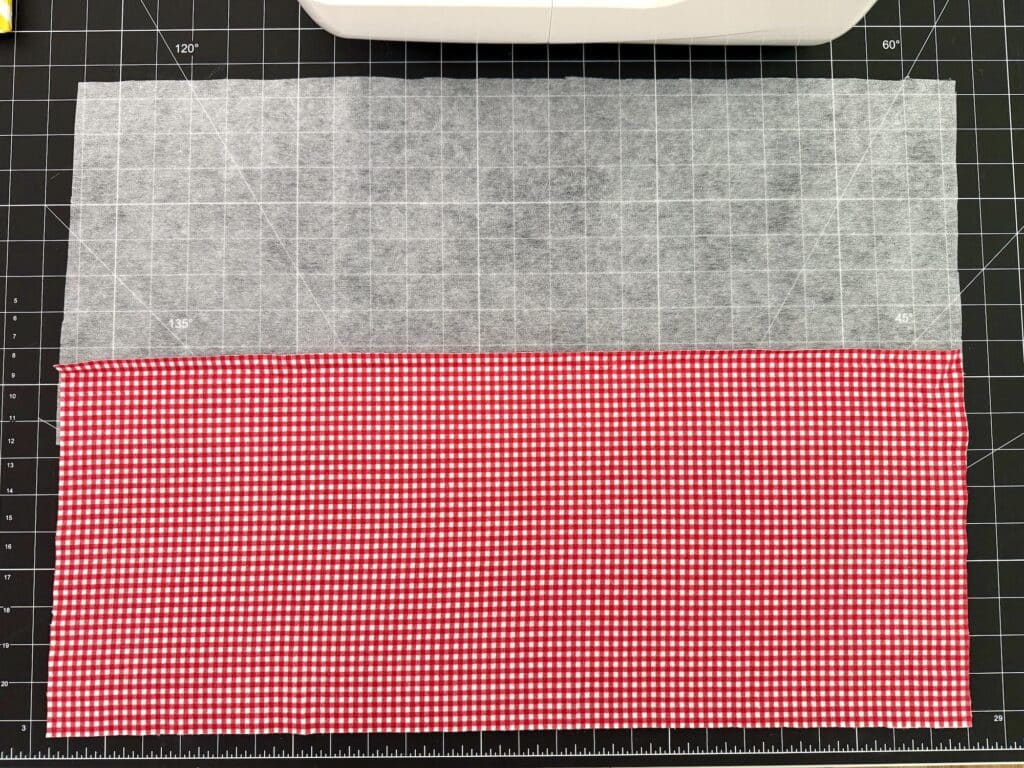 A rectangular piece of red and white checkered fabric is sewn to a piece of grey interfacing on a black grid cutting mat. A sewing machine is partially visible at the top.