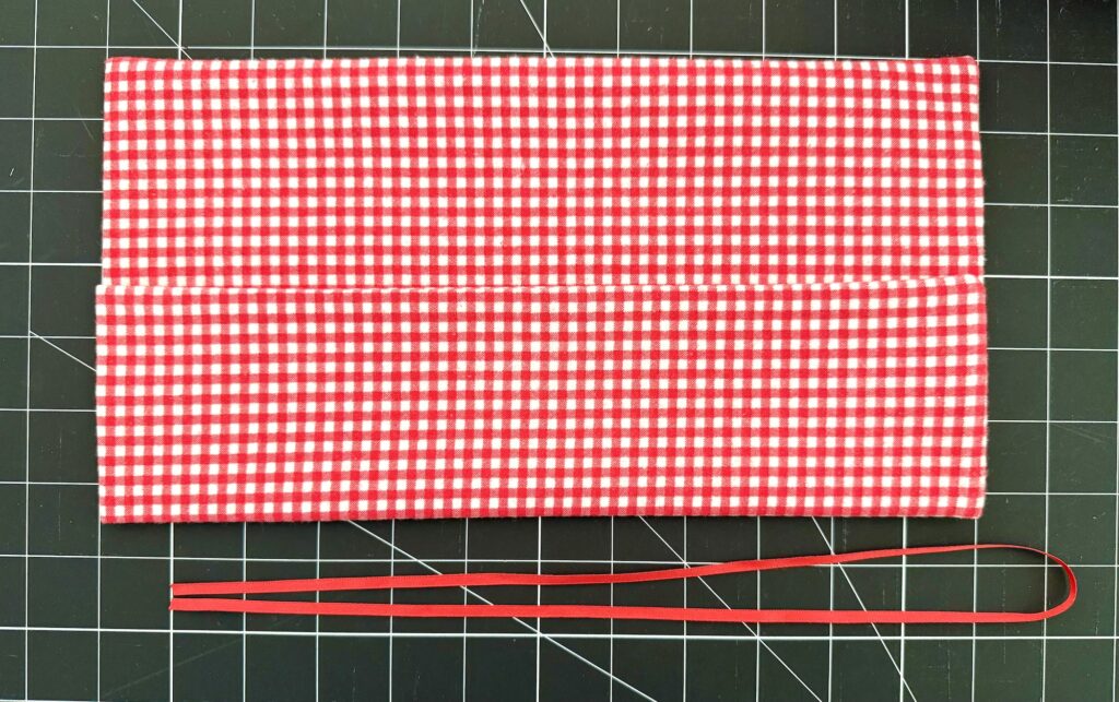 A rectangular piece of red and white checkered fabric is folded in half on a gridded cutting mat, with a red elastic band placed below it.