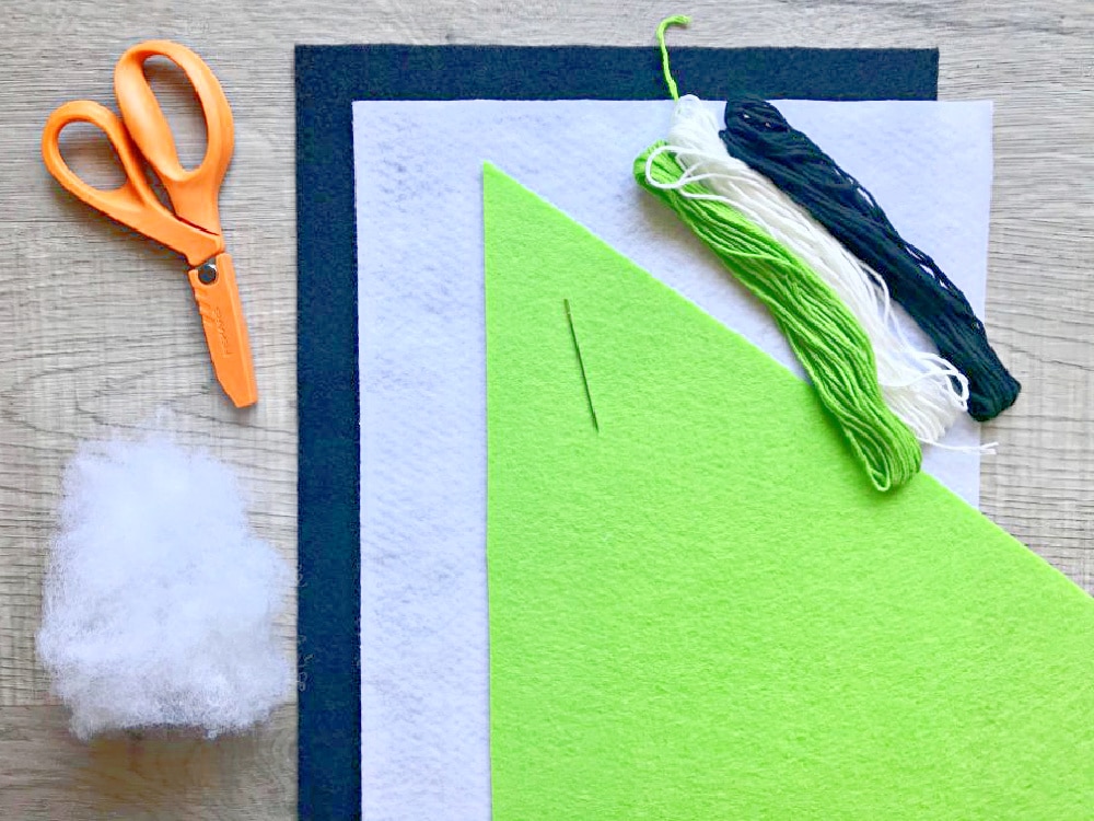 Orange scissors, a sewing needle, green, black, and white embroidery floss, fiberfill stuffing, and sheets of white, black, and green felt are arranged on a wooden surface.