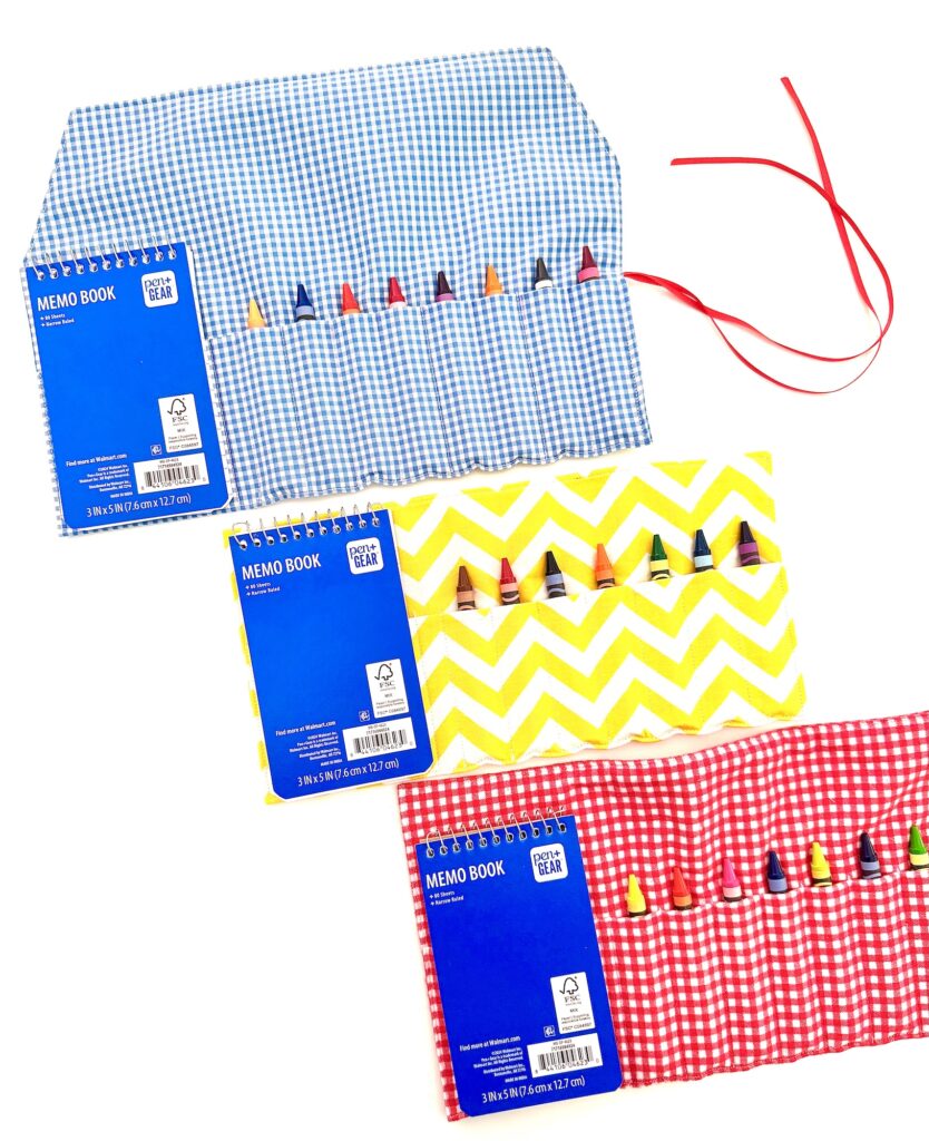 Three colorful crayon roll with notepad cases—blue gingham, red gingham, and yellow chevron—hold multiple crayons and are displayed with open memo books on top.
