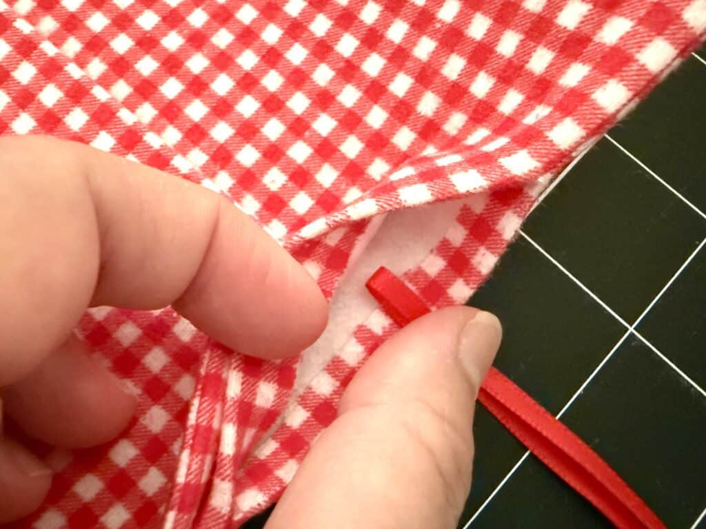 A hand is inserting a red ribbon into the hem of red and white gingham fabric on a black surface with a white grid pattern.
