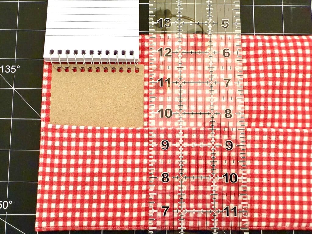 A clear ruler measures a red and white checkered fabric next to a small spiral notepad on a black cutting mat with white gridlines.