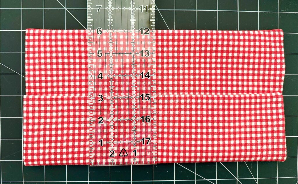 A transparent ruler measuring a folded piece of red and white gingham fabric on top of a black gridded cutting mat.