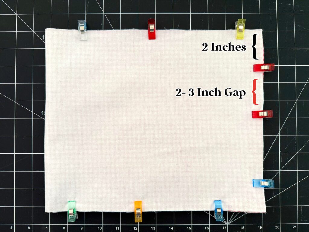 A rectangular white fabric, part of a crayon roll with notepad, is clipped on the edges with colored clips, showing a labeled 2-inch section and a labeled 2–3 inch gap on the right side.