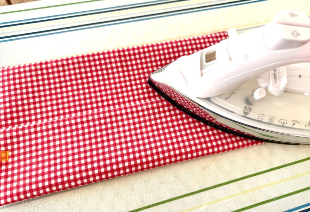 A white iron presses a red and white checkered fabric on a striped ironing board.