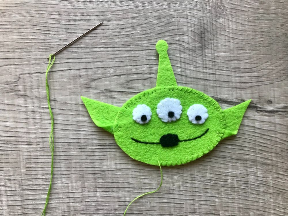 A green felt alien face with three eyes, stitched black mouth, and ears, next to a sewing needle with green thread on a wooden surface.