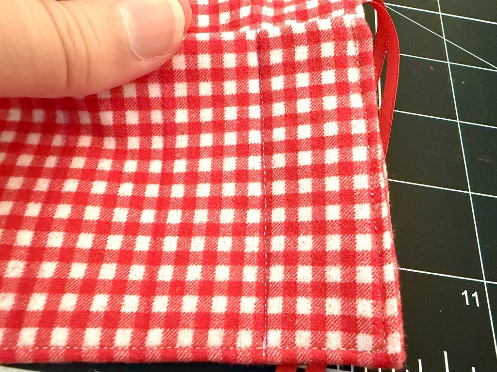 A hand holds a red and white checkered fabric mask with visible stitching beside a black gridded cutting mat, next to a colorful crayon roll with notepad.