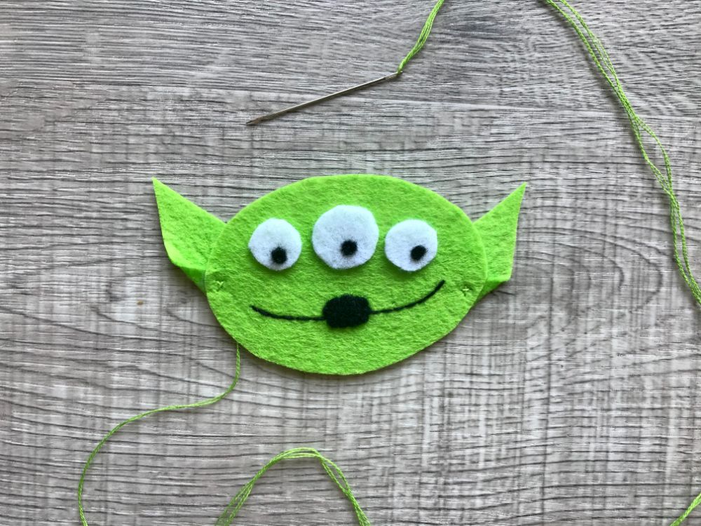 A sewn felt cutout of a green alien face with three eyes, a black nose, and a smiling mouth, along with a needle and green thread on a wooden surface.