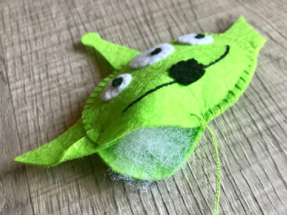 A partially sewn green felt alien head with visible white stuffing and loose thread rests on a wooden surface.