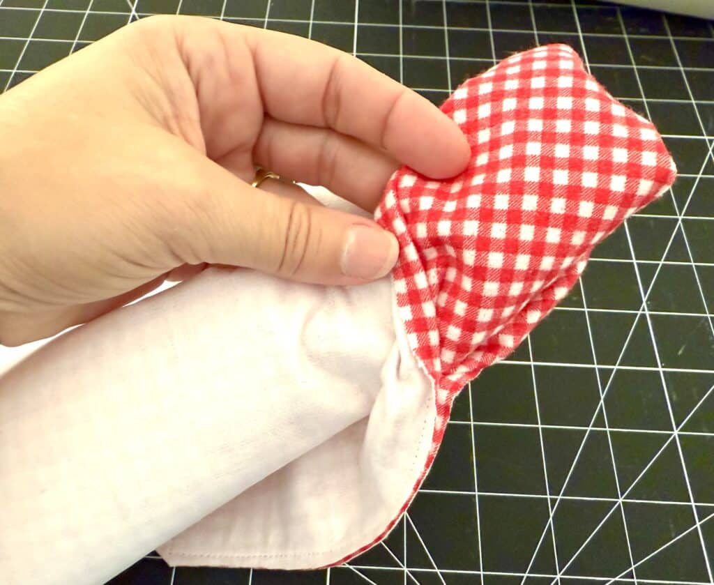 A hand holding the corner of a white fabric item with a red and white checkered section, against a black cutting mat with a grid pattern.