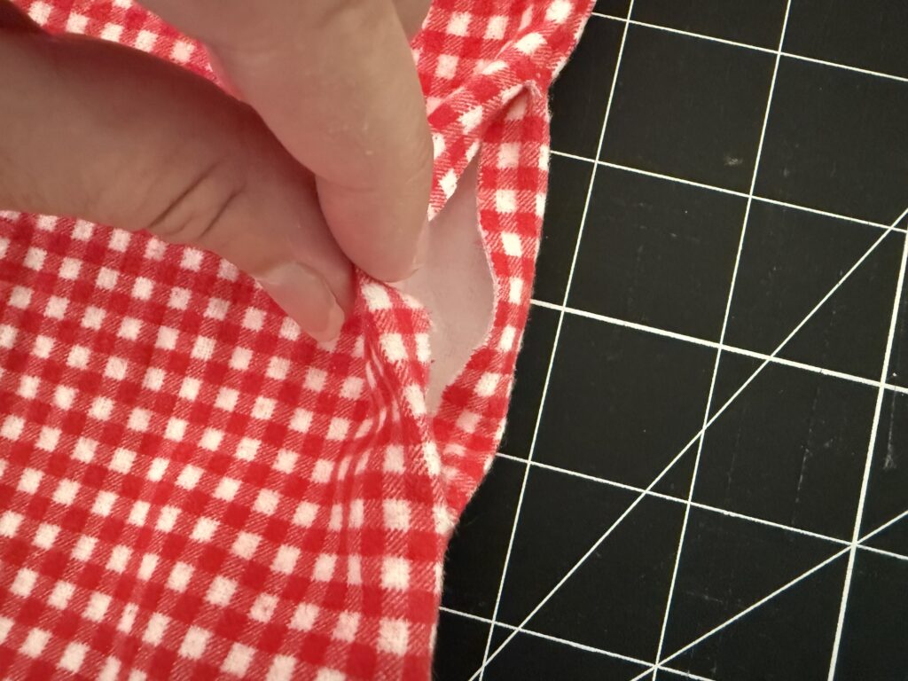 A hand holds open a seam on red and white checkered fabric, revealing a gap—suggesting it's part of a crayon roll with notepad; the fabric rests on a black cutting mat with white grid lines.