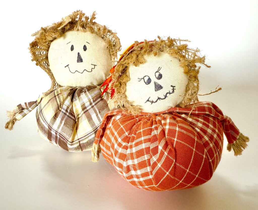 Two handmade scarecrow dolls with stitched faces, straw hair, and plaid fabric bodies—one in brown and yellow, the other in red and white—are placed side by side on a white surface.