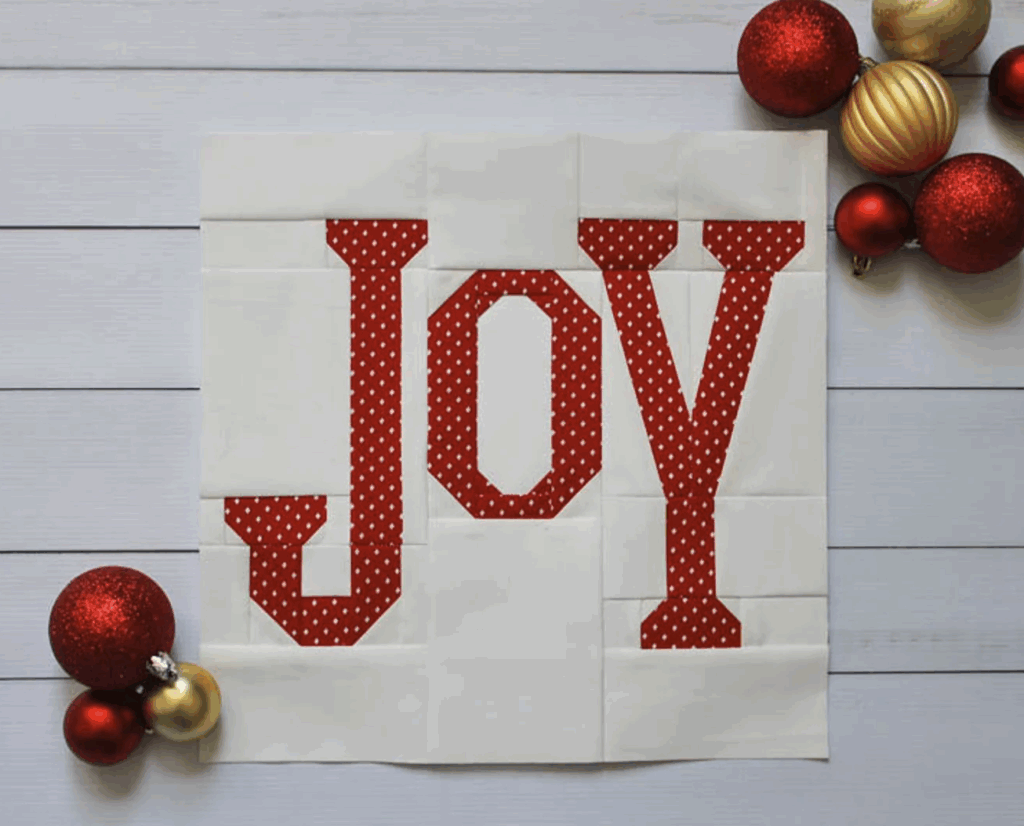 A white quilt square spells "JOY" in red polka dot fabric, surrounded by Christmas quilt blocks and festive red and gold ornaments on a white wooden surface.