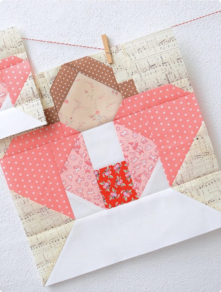 A paper quilt block features a geometric person in a pink and red outfit made of patterned fabrics, clipped with a clothespin against white—an inspiring addition to Christmas quilt blocks.