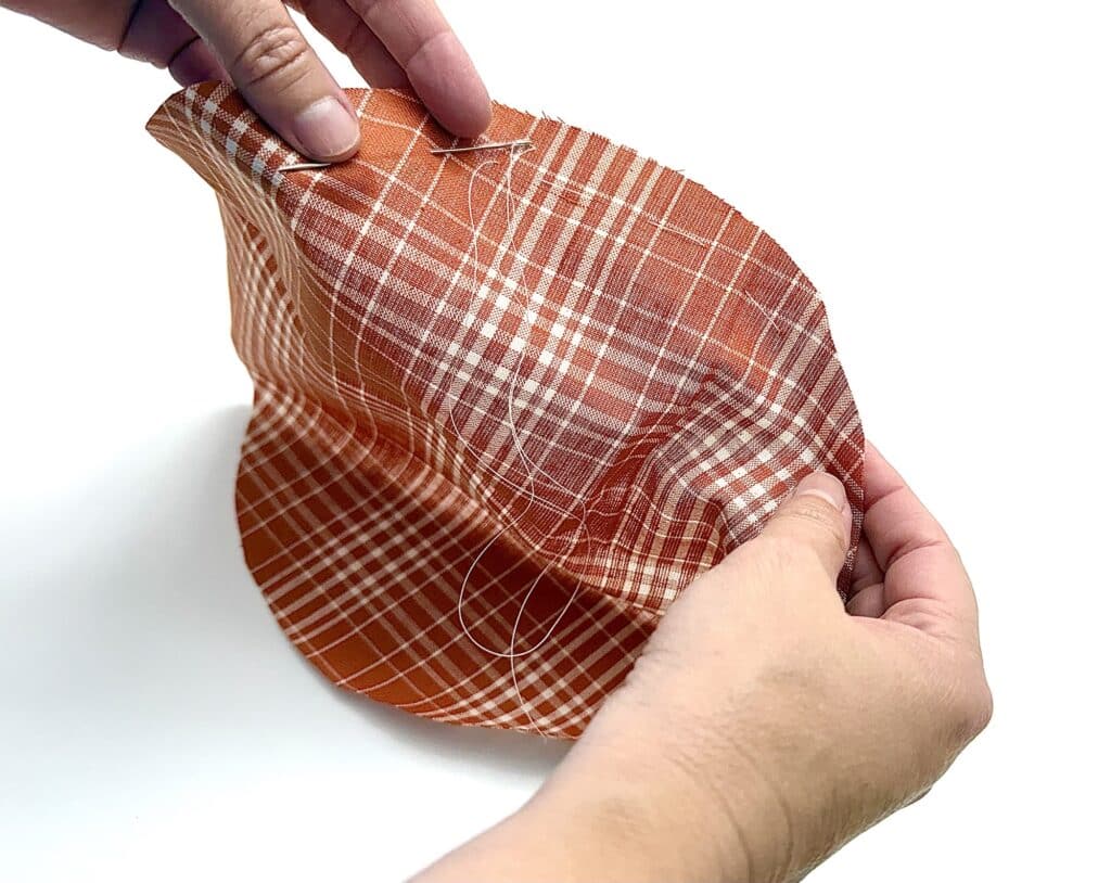 Two hands sewing a curved seam on a piece of orange plaid fabric, with needle and thread visible against a white background.
