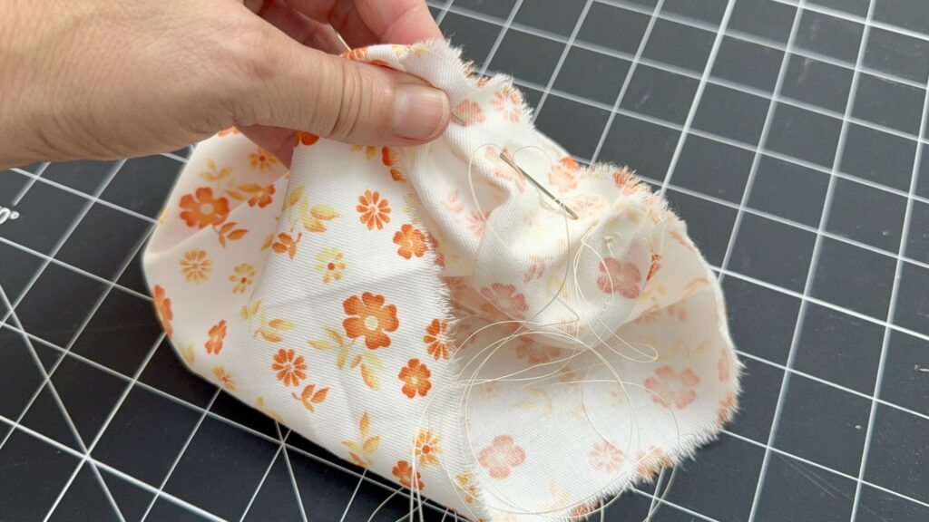 A hand holds a piece of white fabric with orange floral print, sewn with a needle and thread, over a black grid cutting mat.