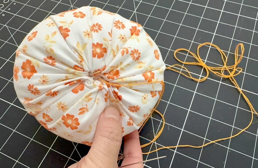 A hand holds a round fabric piece with orange floral print, gathered at the center with orange thread and needle on a black grid cutting mat.