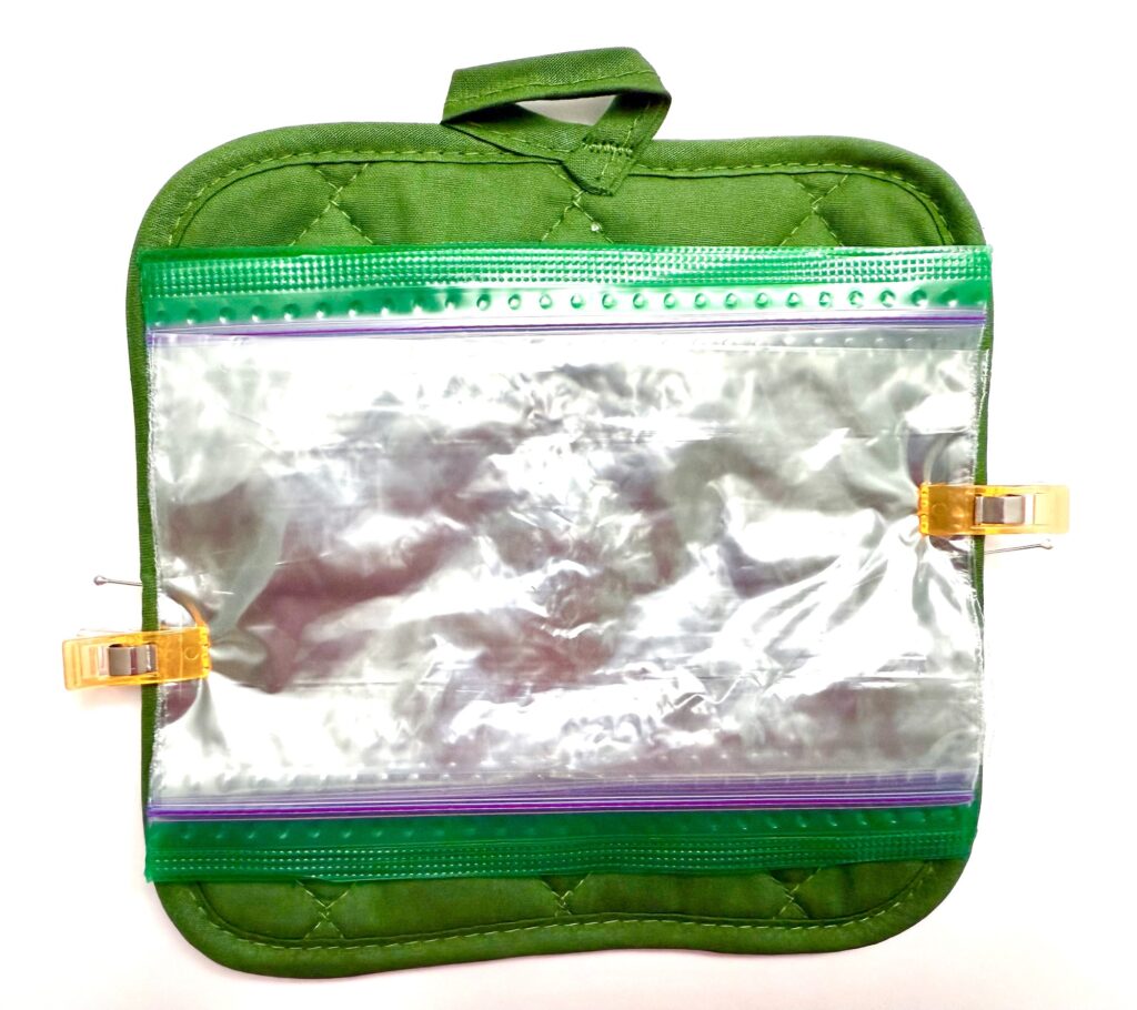 A green quilted pot holder with a clear resealable plastic bag clipped to its front using two yellow binder clips—perfect for keeping small kitchen items handy.