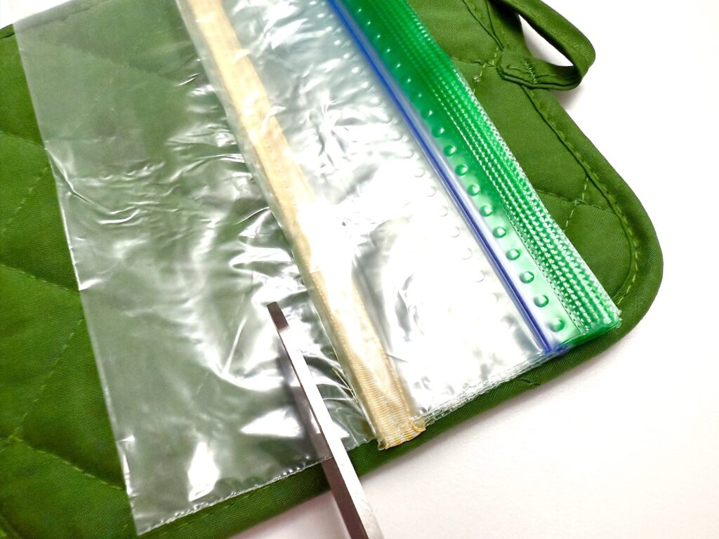 A pair of scissors cutting a row of used zip-top plastic bags sewn onto a green quilted pot holder.