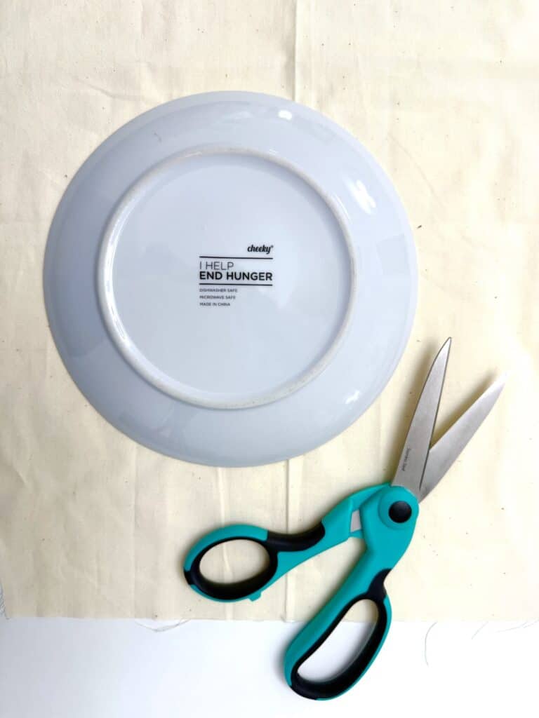 A white plate turned upside down on beige fabric next to a pair of teal and black scissors.