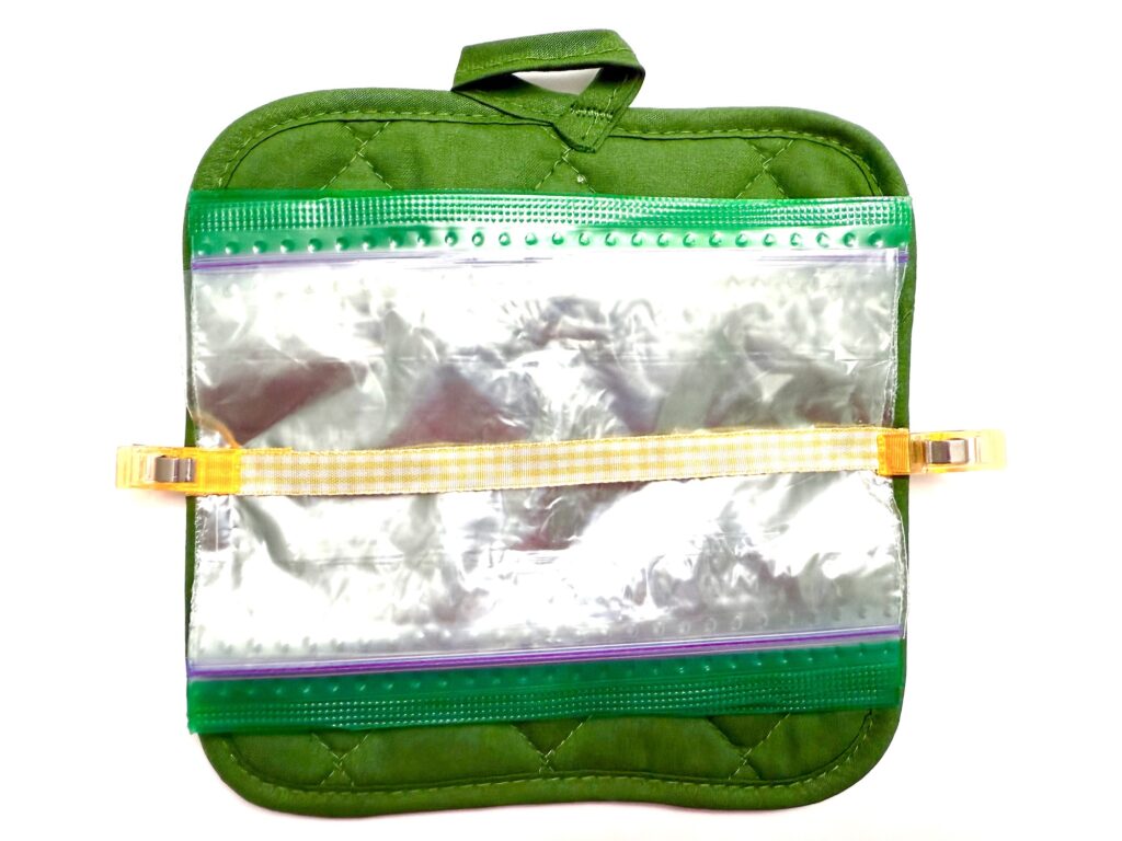 A green quilted pot holder features a clear resealable plastic bag attached to the front with two orange clips and a yellow strap.