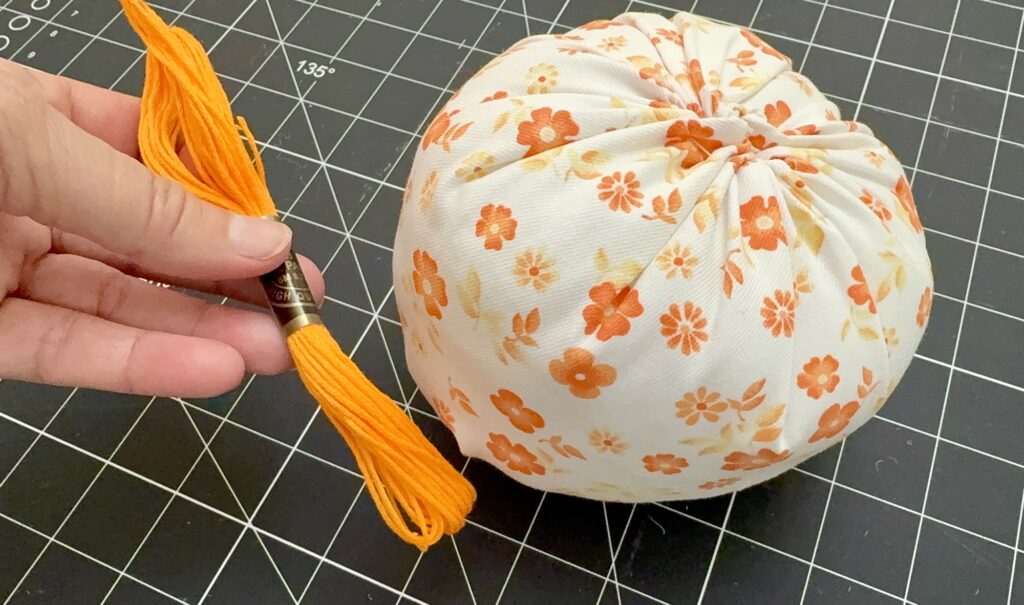 A hand holds orange embroidery floss next to a round fabric pumpkin with an orange floral pattern, placed on a black cutting mat with a grid.