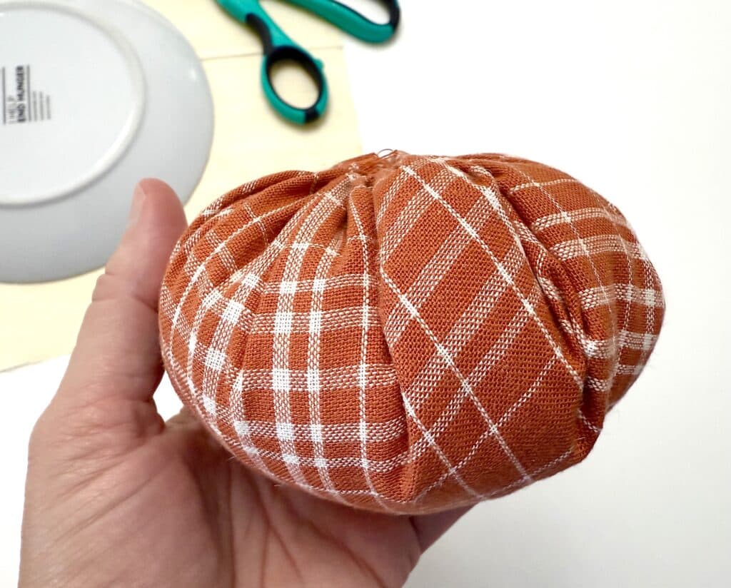 A hand holding a round, orange plaid fabric item, with green-handled scissors and an upside-down plate in the background on a white surface.