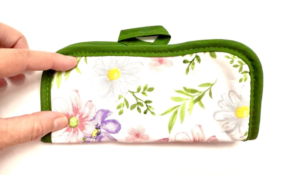A hand holds a floral-patterned, green-edged fabric pot holder pouch with a small handle on top, set against a white background.