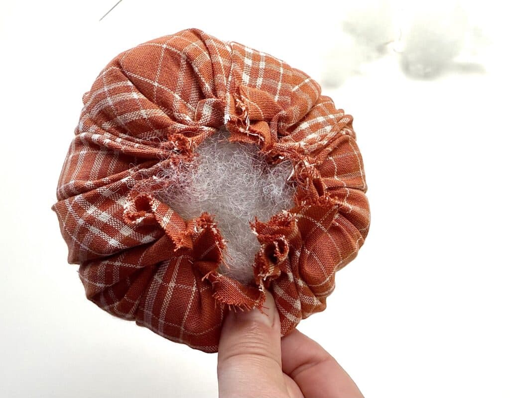 A hand holds a round, orange plaid fabric object with a hole in the center, exposing white stuffing inside against a white background.