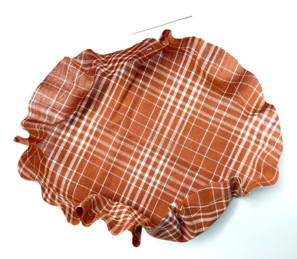 A round piece of orange plaid fabric with gathered edges, placed next to a sewing needle on a white surface.