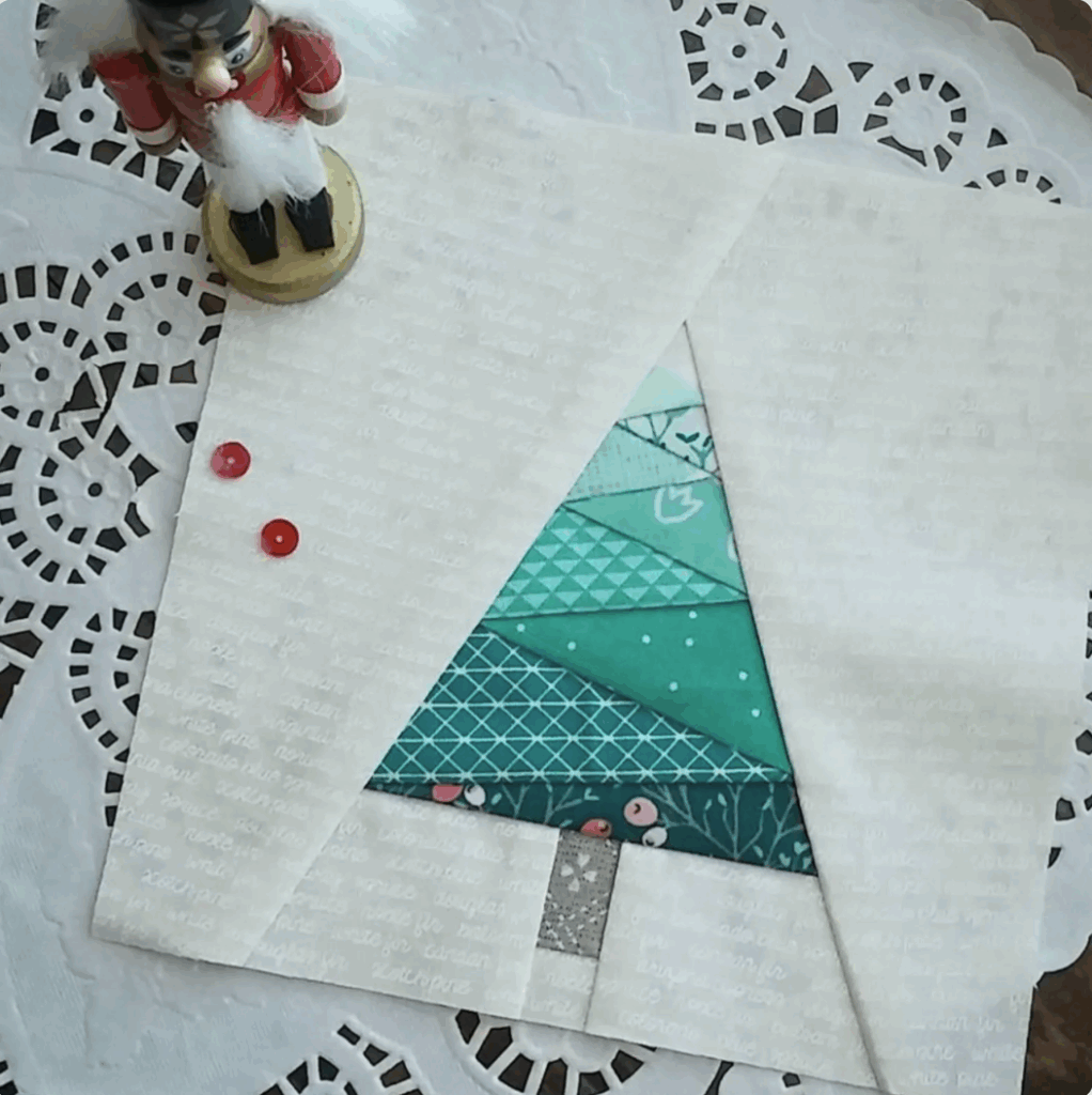 A paper-pieced Christmas quilt block features a teal geometric tree on a white background, next to a nutcracker figurine and two red sequins, all displayed on a lace doily.
