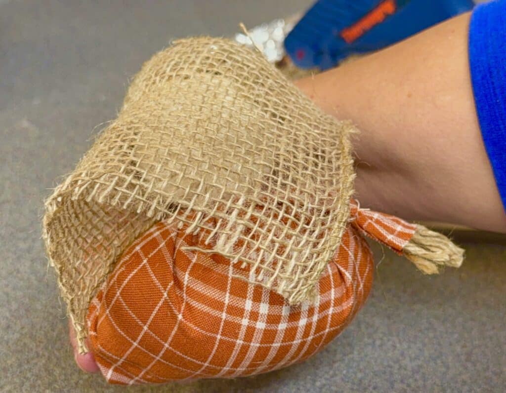 A hand holding a fabric pumpkin with an orange checkered pattern and a piece of burlap covering the top.