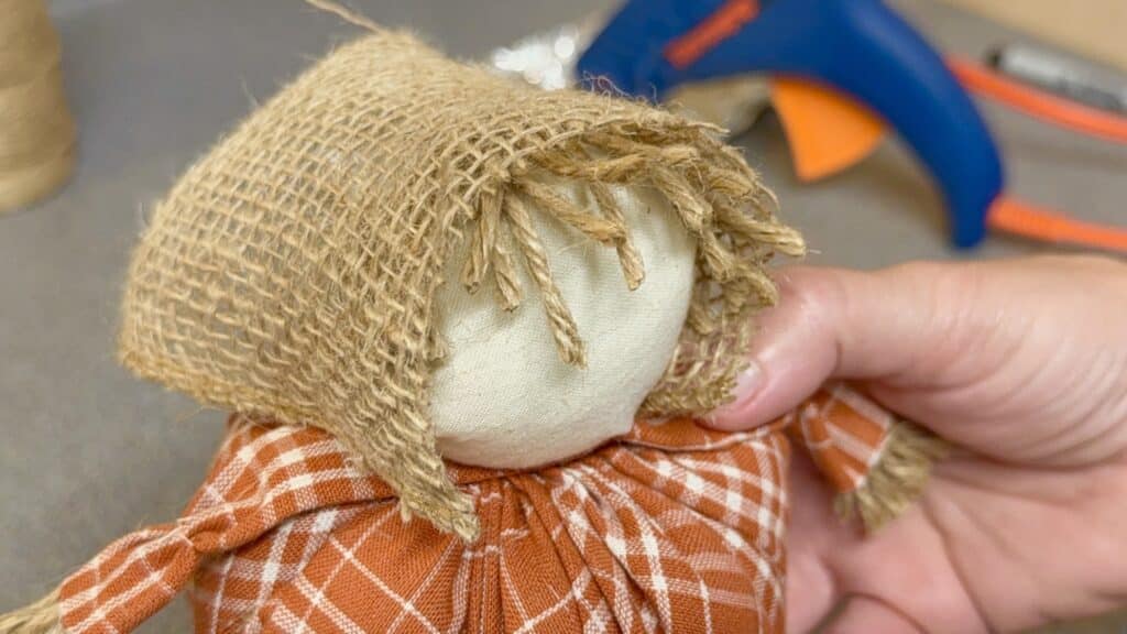 A hand holds a small homemade doll with a burlap hood and orange plaid dress; a hot glue gun is visible in the background.