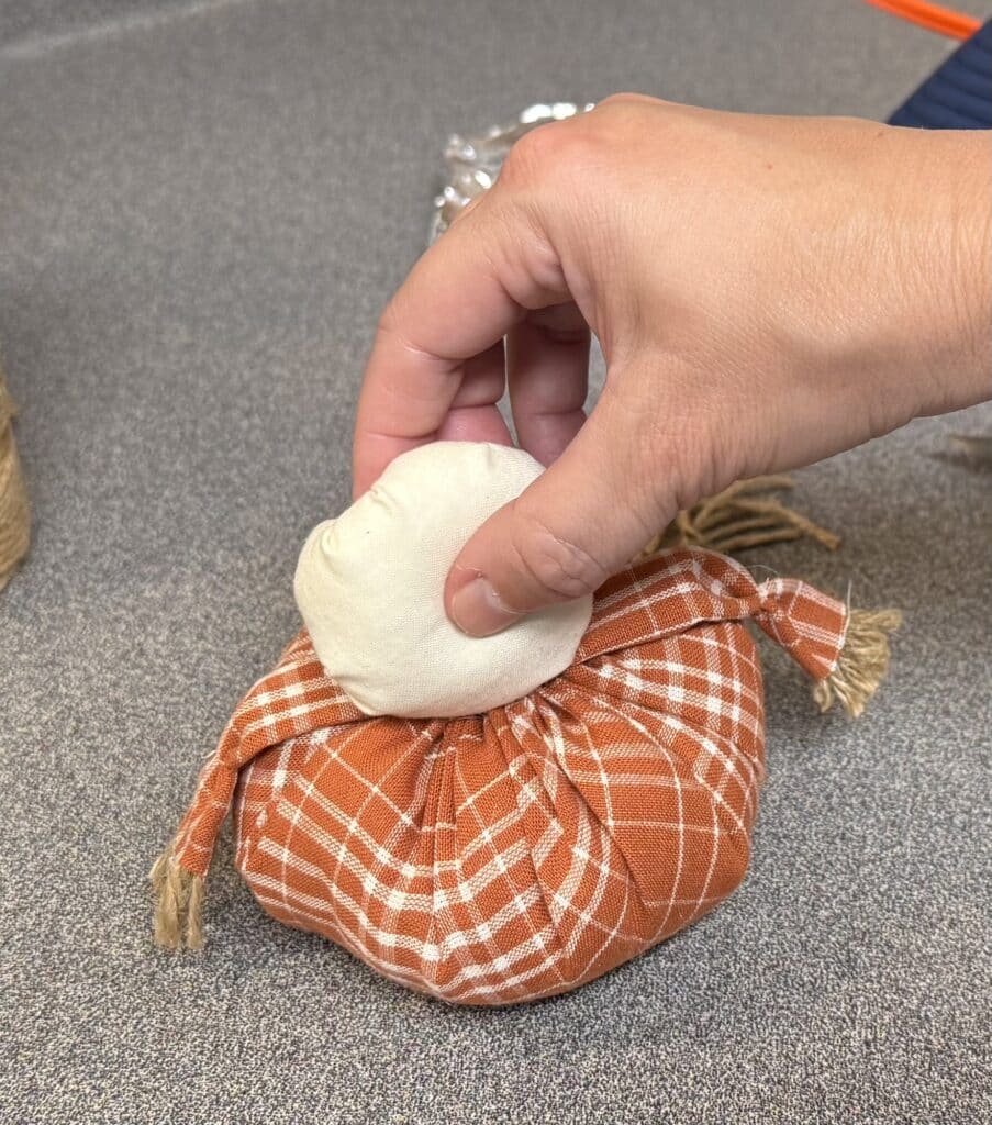 A hand is placing a small round fabric head onto a pumpkin-shaped object wrapped in orange plaid cloth, likely as part of a craft project.