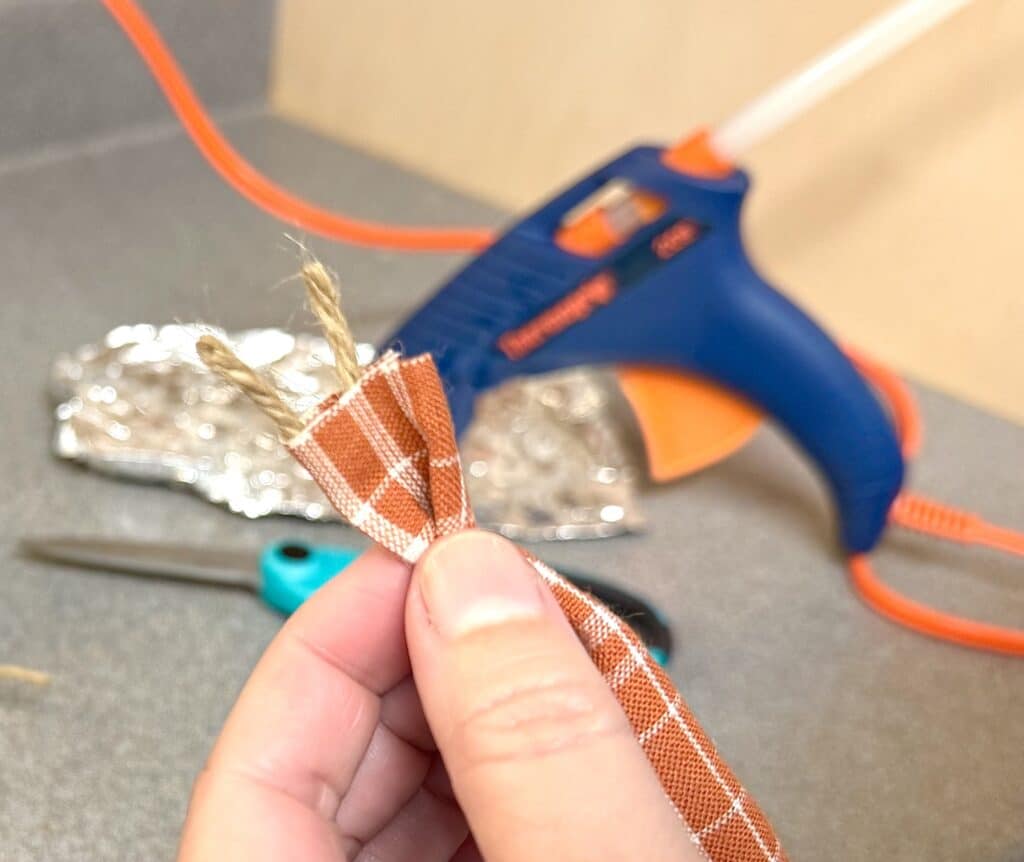 A hand holds a piece of plaid fabric and twine; a hot glue gun, scissors, and foil are on the counter in the background.